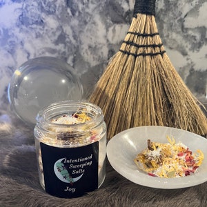 May include: A glass jar filled with white bath salts, herbs, and flower petals. The jar has a black label that reads "Intentioned Sweeping Salts Joy".