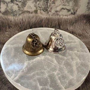 May include: Two small bells with pentagram charms, one gold and one silver, on a white selenite charging plate.