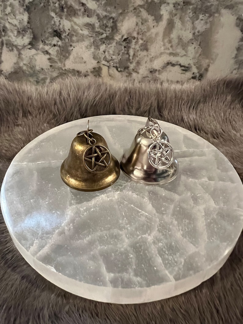 May include: Two small bells with pentagram charms, one gold and one silver, on a white selenite charging plate.