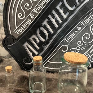 Puede incluir: Tres botellas de vidrio con tapones de corcho. Las botellas est&aacute;n vac&iacute;as y son transparentes. Las botellas est&aacute;n sobre una superficie marr&oacute;n y peluda. Un letrero negro con letras blancas dice "Apothecary" con las palabras "Potions & Poisons" y "Tonics & Tinctures" a cada lado.