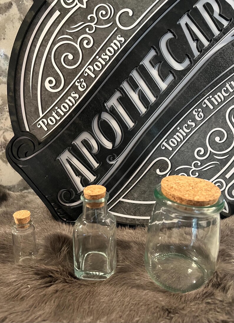 Puede incluir: Tres botellas de vidrio con tapones de corcho. Las botellas est&aacute;n vac&iacute;as y son transparentes. Las botellas est&aacute;n sobre una superficie marr&oacute;n y peluda. Un letrero negro con letras blancas dice "Apothecary" con las palabras "Potions & Poisons" y "Tonics & Tinctures" a cada lado.