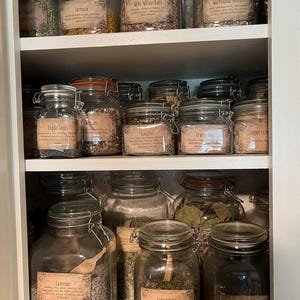Puede incluir: Un estante de armario blanco lleno de frascos de vidrio que contienen hierbas y flores secas. Los frascos tienen etiquetas con los nombres de las hierbas y flores, como "Lavanda", "Menta", "Tr&eacute;bol rojo", "Ajenjo", "Corteza de sauce blanco" y "Semillas de amapola".