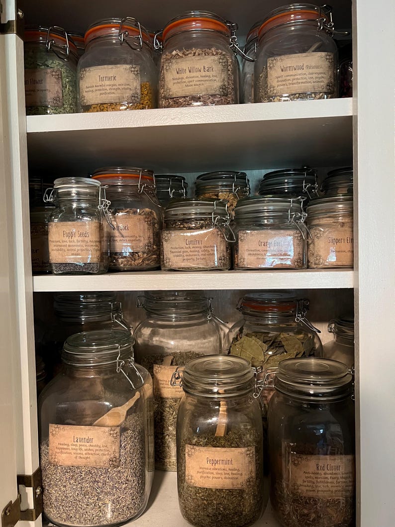 Puede incluir: Un estante de armario blanco lleno de frascos de vidrio que contienen hierbas y flores secas. Los frascos tienen etiquetas con los nombres de las hierbas y flores, como "Lavanda", "Menta", "Tr&eacute;bol rojo", "Ajenjo", "Corteza de sauce blanco" y "Semillas de amapola".