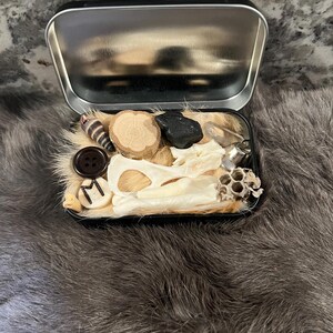 May include: A small metal tin filled with various objects, including a bone, a button, a piece of wood, a black rock, and a honeycomb. The tin is open and the objects are visible.