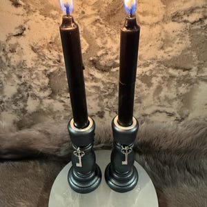 Könnte beinhalten: Zwei schwarze Kerzen mit hellen Flammen, in dekorativen schwarzen Haltern mit Schlüsselanhängern. Die Kerzen stehen auf einer weißen, runden Basis, mit strukturiertem Hintergrund.