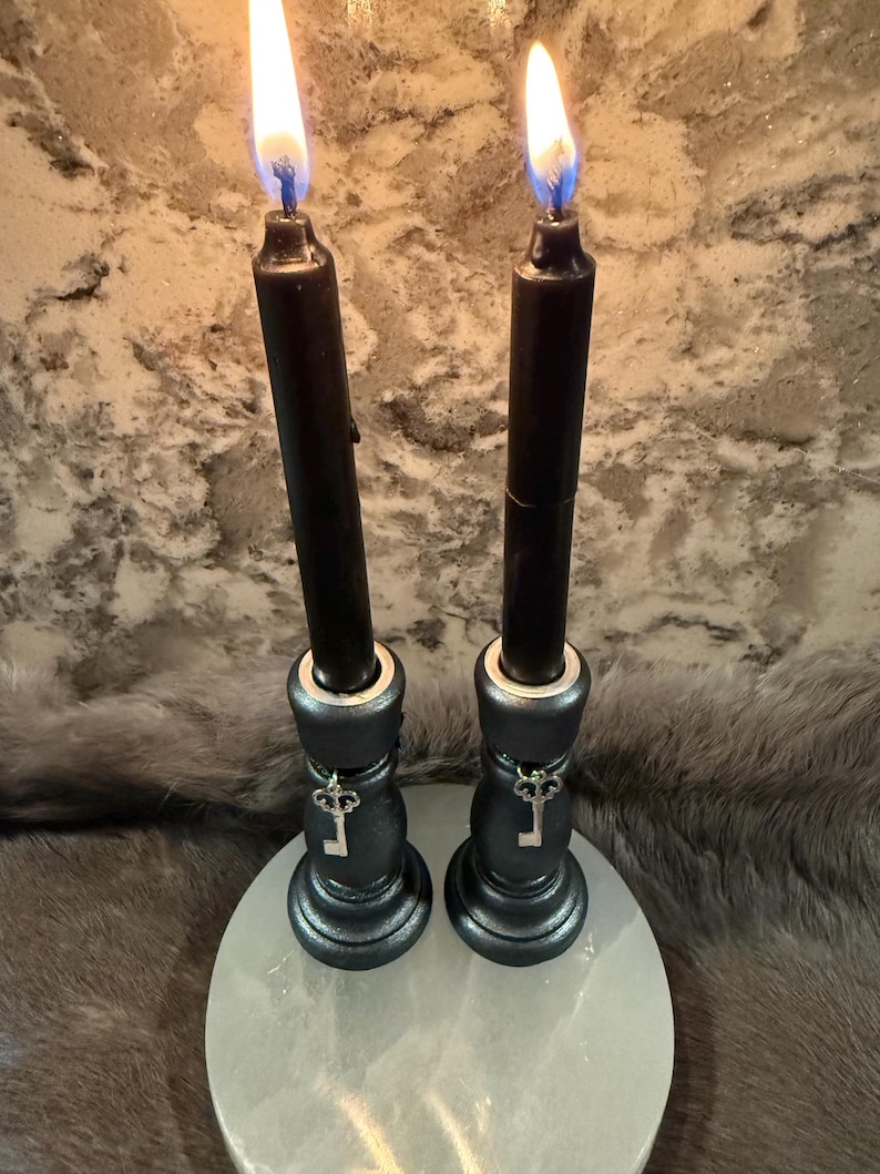 May include: Two black candles with bright flames, set in decorative black holders with key charms. The candles are on a white, round base, with a textured background.