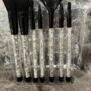 Puede incluir: Conjunto de siete pinceles de maquillaje negros con mangos transparentes rellenos de purpurina plateada. Los pinceles están en una caja de plástico transparente con un fondo de purpurina plateada.