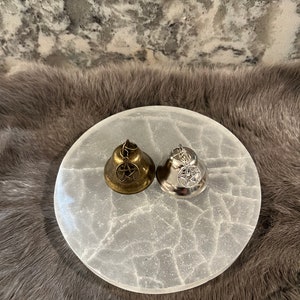May include: Two small bells with pentagram charms. One bell is silver and the other is gold. The bells are sitting on a white selenite charging plate.