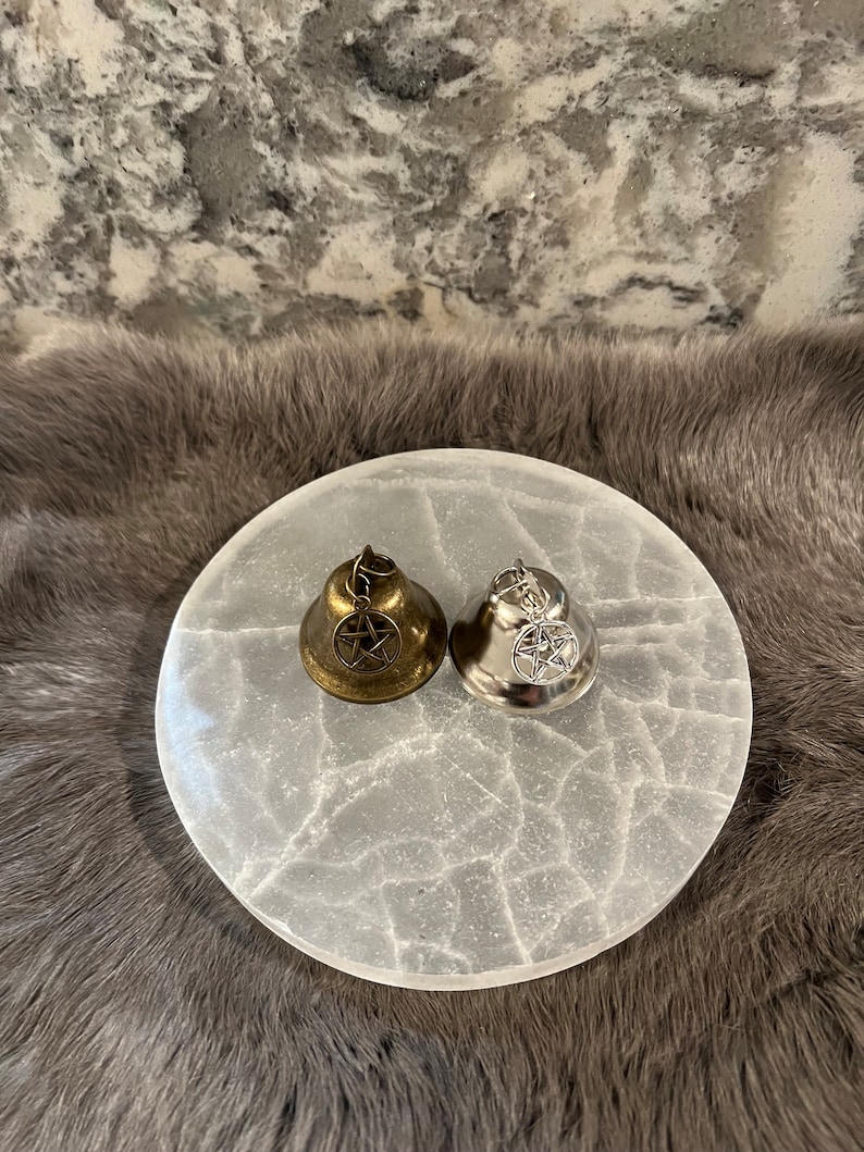 May include: Two small bells with pentagram charms. One bell is silver and the other is gold. The bells are sitting on a white selenite charging plate.
