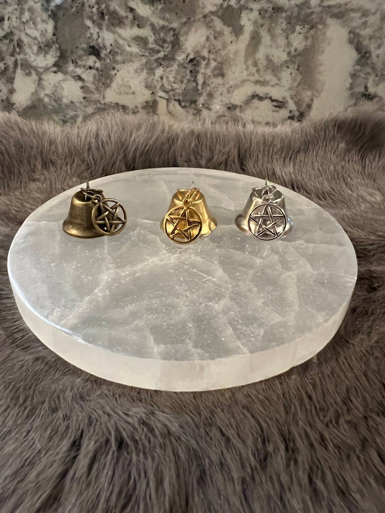 Pu&ograve; includere: Tre piccole campane d'argento con un ciondolo a pentagramma appeso a ciascuna. Le campane sono appoggiate su un piatto di ricarica in selenite rotondo e bianco.
