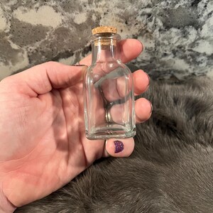 Puede incluir: Una peque&ntilde;a botella de vidrio transparente con un tap&oacute;n de corcho. La botella est&aacute; vac&iacute;a y tiene una forma cuadrada.