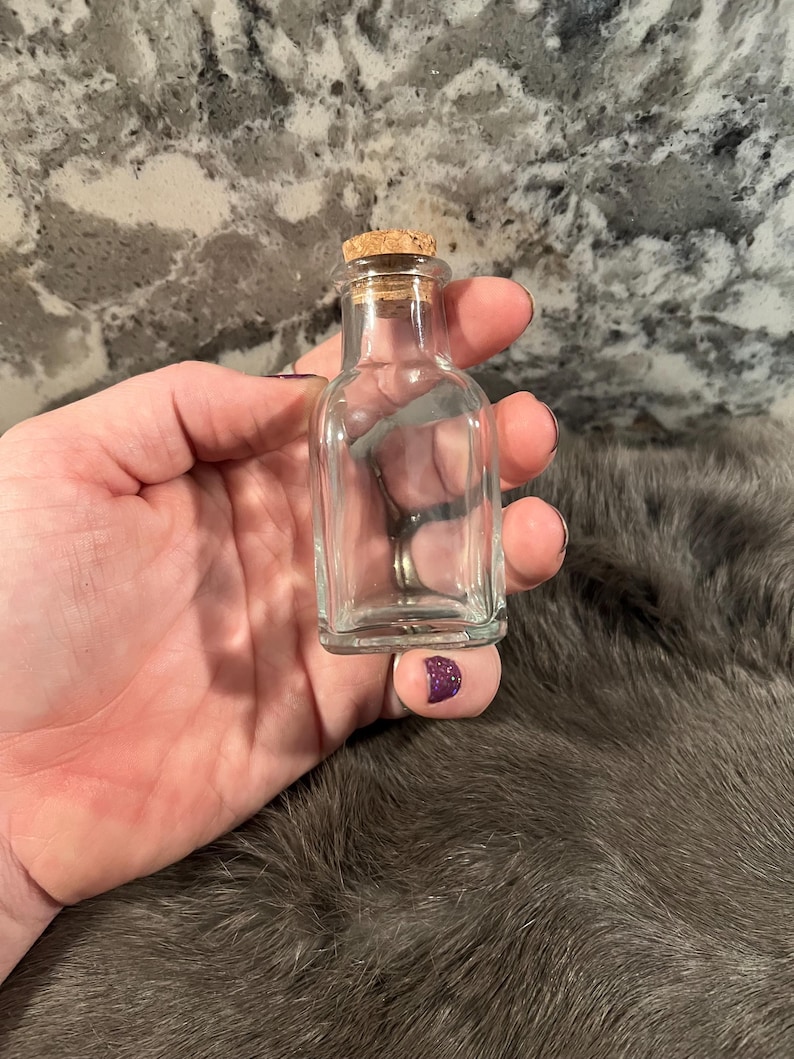 Puede incluir: Una peque&ntilde;a botella de vidrio transparente con un tap&oacute;n de corcho. La botella est&aacute; vac&iacute;a y tiene una forma cuadrada.