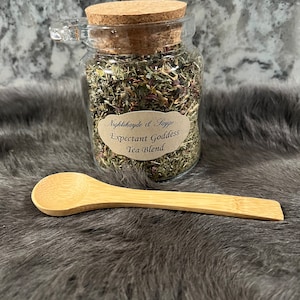 Puede incluir: Un frasco de vidrio con tapa de corcho lleno de hierbas secas y una etiqueta que dice "Nightshade & Sage Expectant Goddess Tea Blend". Una cuchara de madera está al lado del frasco.