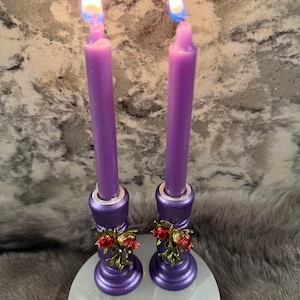 May include: Two lit purple taper candles in decorative holders. The holders are purple with silver accents and adorned with red and green floral embellishments. The candles are on a white, round surface.