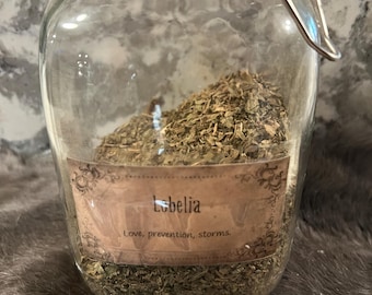 Könnte beinhalten: Ein klares Glas mit Metallverschluss und orangefarbenem Deckel, gefüllt mit getrockneten grünen Kräutern. Auf dem Etikett des Glases steht "Lobelia" mit den Worten "Love, prevention, storms". Das Glas steht vor einem strukturierten Hintergrund.