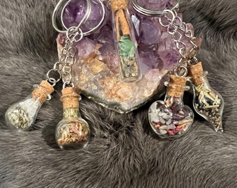 May include: Five small glass bottles with cork tops filled with various herbs, crystals, and other natural elements. The bottles are attached to a silver chain and key ring. The bottles are sitting on a bed of amethyst crystals.