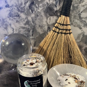 May include: A glass jar filled with white bath salts, herbs, and crystals. The jar has a black label that reads "Intentioned Sweeping Salts Energy Cleansing".