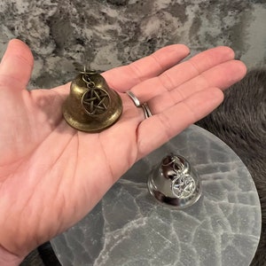 May include: Two small bells with a pentagram charm. The bell on the left is gold and the bell on the right is silver.