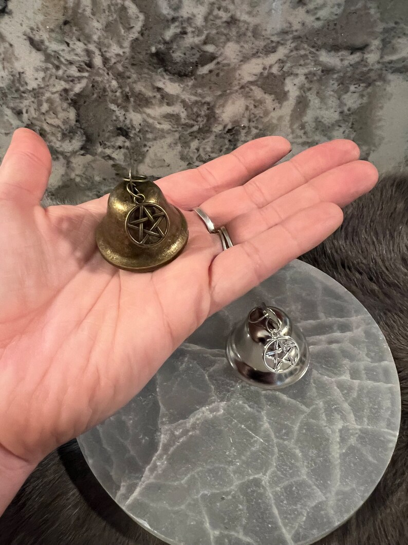 May include: Two small bells with a pentagram charm. The bell on the left is gold and the bell on the right is silver.
