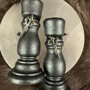 May include: Two black metallic candle holders with key charms. The candle holders have a textured finish and are decorated with a silver key charm. The candle holders are on a white circular surface.