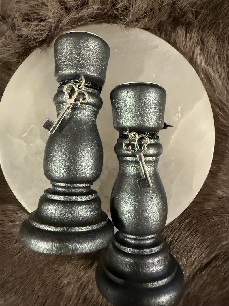 May include: Two black metallic candle holders with key charms. The candle holders have a textured finish and are decorated with a silver key charm. The candle holders are on a white circular surface.