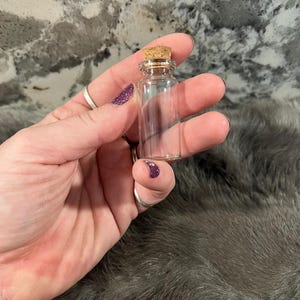 Puede incluir: Una peque&ntilde;a botella de vidrio transparente con un tap&oacute;n de corcho. La botella est&aacute; vac&iacute;a y parece estar hecha de vidrio grueso.