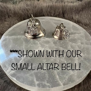 May include: Two silver bells with a pentagram charm hanging from each. The bells are sitting on a white selenite altar plate. The text "**** SHOWN WITH OUR SMALL ALTAR BELL!" is written on the plate.