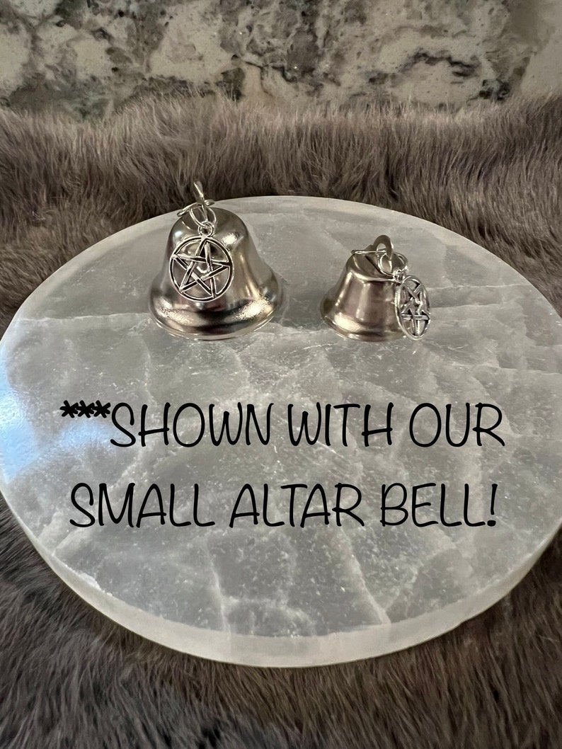 May include: Two silver bells with a pentagram charm hanging from each. The bells are sitting on a white selenite altar plate. The text "**** SHOWN WITH OUR SMALL ALTAR BELL!" is written on the plate.