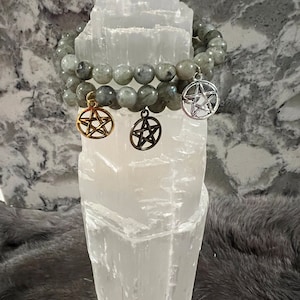 May include: Two grey gemstone bracelets with silver and gold pentagram charms. The bracelets are displayed on a white selenite crystal.