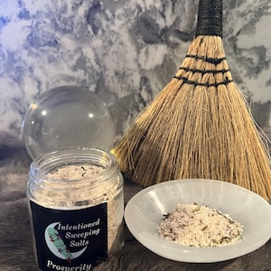 May include: A glass jar of white and green sweeping salts with a black label that reads "Intentioned Sweeping Salts Prosperity". The jar is sitting on a gray fuzzy surface next to a white dish with more of the salts and a brown broom.