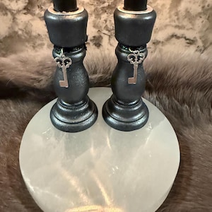 May include: Two black candles sit atop dark gray candle holders with key charms, placed on a white, round, stone base. The candle holders have a metallic finish. The background is a blurred, textured pattern.