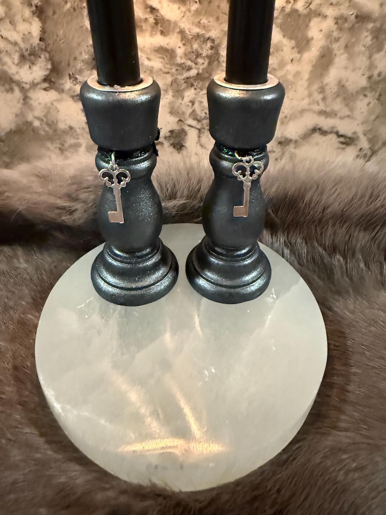 May include: Two black candles sit atop dark gray candle holders with key charms, placed on a white, round, stone base. The candle holders have a metallic finish. The background is a blurred, textured pattern.