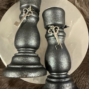 May include: Two dark gray candle holders with a metallic finish. Each holder has a key charm attached with black string. The holders are on a white, round surface, with a brown, furry background.