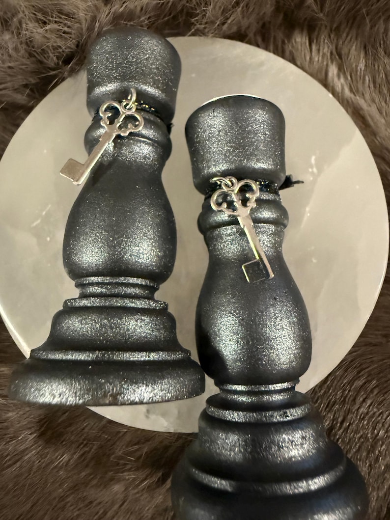 May include: Two dark gray candle holders with a metallic finish. Each holder has a key charm attached with black string. The holders are on a white, round surface, with a brown, furry background.