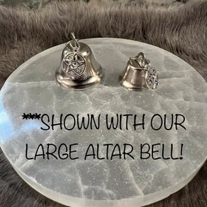 Pu&ograve; includere: Due campane d'argento con un ciondolo a pentagramma su un piatto da altare in selenite bianca. Il testo "**** SHOWN WITH OUR LARGE ALTAR BELL!" &egrave; stampato sul piatto.