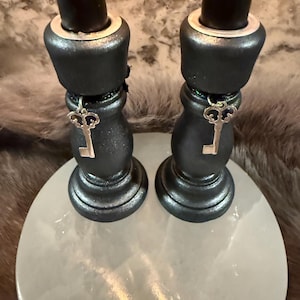May include: Two black candle holders with silver key charms hanging from the base. The candle holders sit on a round, white, stone-like surface. The candle holders have a metallic sheen.