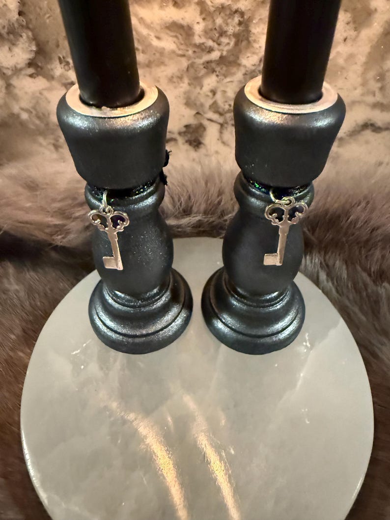 May include: Two black candle holders with silver key charms hanging from the base. The candle holders sit on a round, white, stone-like surface. The candle holders have a metallic sheen.