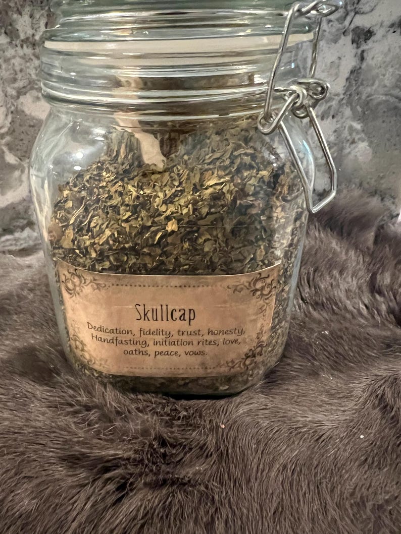 Puede incluir: Un frasco de vidrio lleno de hierbas secas etiquetado como "Skullcap". La etiqueta enumera los usos de la hierba, que incluyen dedicaci&oacute;n, fidelidad, confianza, honestidad, uni&oacute;n de manos, ritos de iniciaci&oacute;n, amor, juramentos, paz y votos.