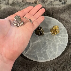 Pu&ograve; includere: Tre campane d'argento, oro e nero con un design a pentagramma. Le campane sono su una superficie rotonda, testurizzata e bianca.