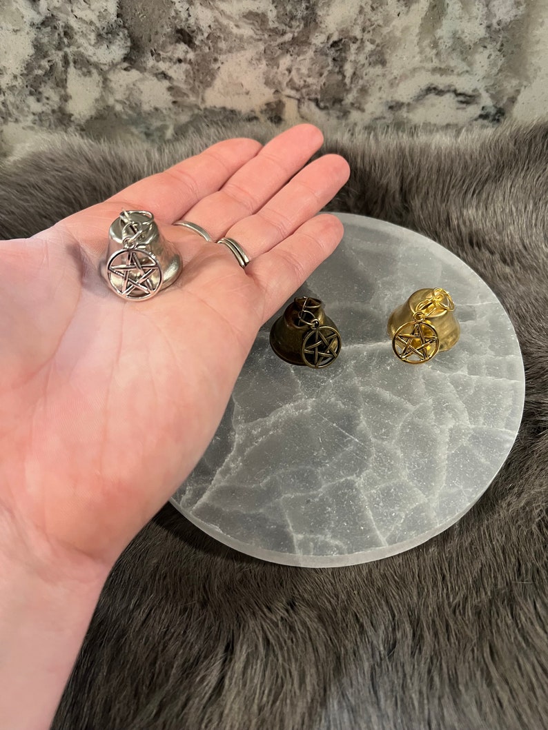 Pu&ograve; includere: Tre campane d'argento, oro e nero con un design a pentagramma. Le campane sono su una superficie rotonda, testurizzata e bianca.