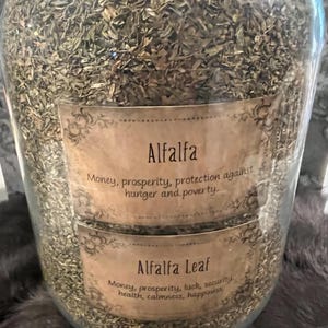 Puede incluir: Un gran tarro de vidrio lleno de hojas de alfalfa secas. Se ven dos etiquetas, una que dice "Alfalfa" y otra "Alfalfa Leaf". Las etiquetas enumeran propiedades asociadas como dinero, prosperidad y protección.