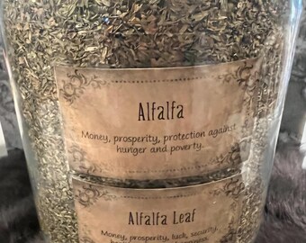 Könnte beinhalten: Ein großes Glasgefäß gefüllt mit getrockneten Alfalfa-Blättern. Zwei Etiketten sind sichtbar, eines mit der Aufschrift "Alfalfa" und das andere mit der Aufschrift "Alfalfa Leaf". Die Etiketten listen zugeordnete Eigenschaften wie Geld, Wohlstand und Schutz auf.