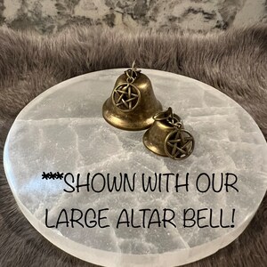 Pu&ograve; includere: Due campane dorate con un designo a pentagramma su una superficie rotonda bianca. Il testo "***SHOWN WITH OUR LARGE ALTAR BELL!" &egrave; scritto sulla superficie.