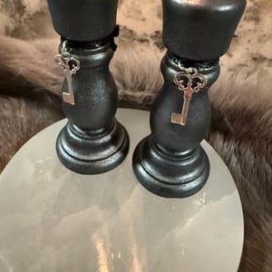 May include: Two black candle holders with silver key charms, resting on a white, round surface. The candle holders have a metallic finish and are adorned with decorative key pendants. The background features a textured, neutral-toned surface.