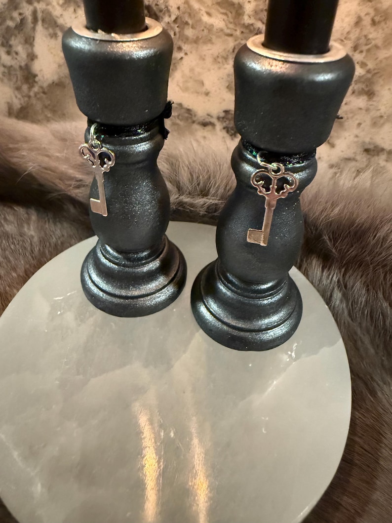 May include: Two black candle holders with silver key charms, resting on a white, round surface. The candle holders have a metallic finish and are adorned with decorative key pendants. The background features a textured, neutral-toned surface.