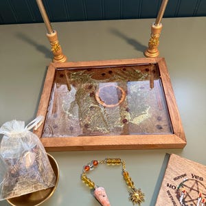 May include: A wooden divination board with a decorative frame, two gold candle holders, a pendulum, a small bag, and a wooden coaster with text. The board has a clear surface with embedded decorative elements.