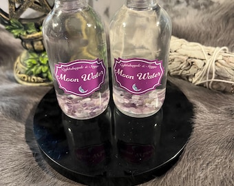 May include: Two clear glass bottles with black lids and labels that say "Moon Water" with purple crystals inside. The bottles are on a black circular stand.
