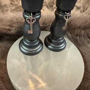 May include: Two black candle holders with silver key charms hanging from them. The candle holders are on a white, round surface. The background is a blurred, furry texture.