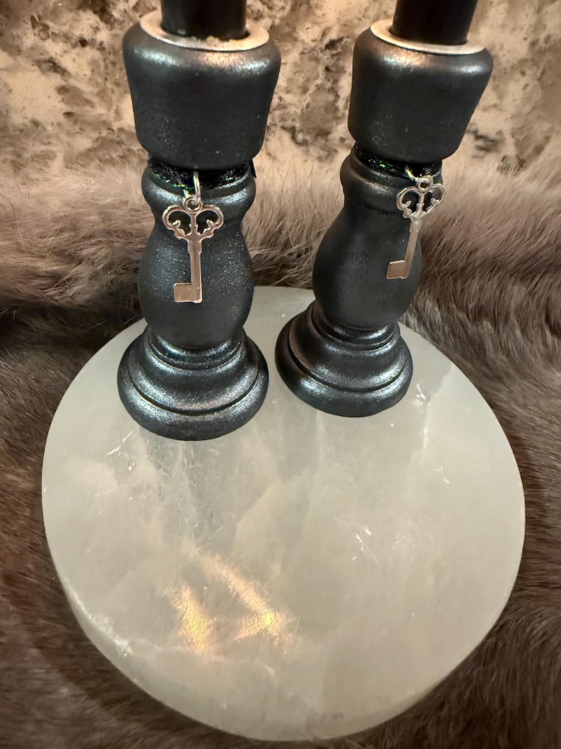 May include: Two black candle holders with silver key charms hanging from them. The candle holders are on a white, round surface. The background is a blurred, furry texture.