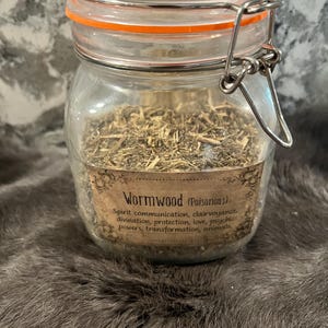 Puede incluir: Un frasco de vidrio con tapa de cierre metálico lleno de hierbas secas etiquetadas como "Wormwood (Poisonous)". La etiqueta enumera los usos de la hierba, incluyendo la comunicación espiritual, la clarividencia, la adivinación, la protección, el amor, los poderes psíquicos, la transformación y los animales.